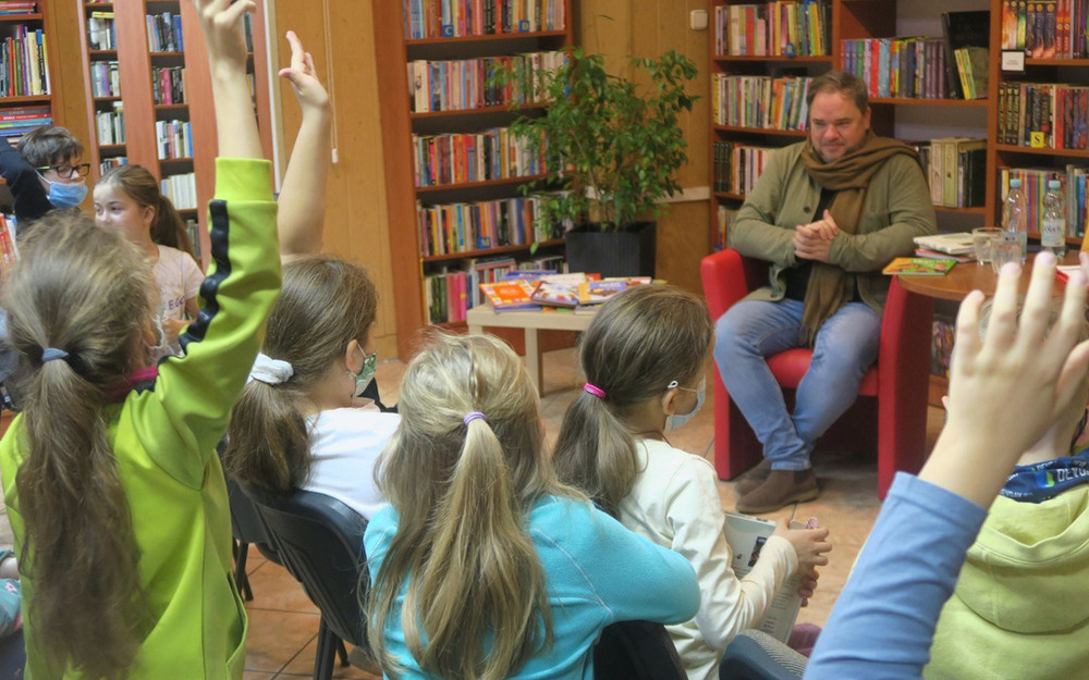 Grzegorz Kasdepke siedzący na tle regałów z książkami oraz dzieci siedzące naprzeciwko autora z podniesionymi rękoma i zadające pytania 
